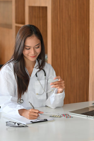 Young asian female pharmacist holding a pack of pills, tablet blisters, medicine and writing a drugs prescription on paperの写真素材