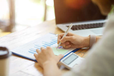 Close-up Businesswoman write to do list on post note, working on financial data chart report and laptop computer at her deskの写真素材