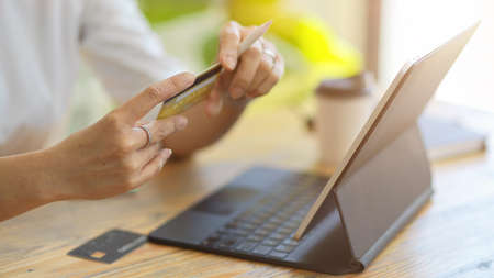 Young adult woman holding credit card, looking at her credit card number. Tablet and credit card on table. Close-upの写真素材
