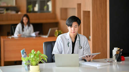 A young male doctor is using a laptop computer. In the hospital office, a male physician reads a surgery plan and conducts medical research on laptopの写真素材