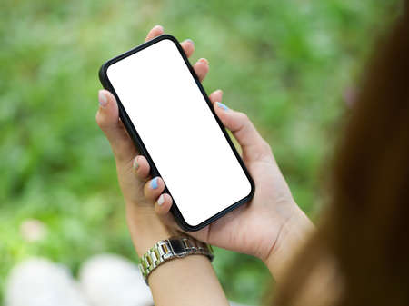 Close-up of a female holding a smartphone with a blank screen mockup for montage against a blurred green grass background. Concept of device equipmentの写真素材
