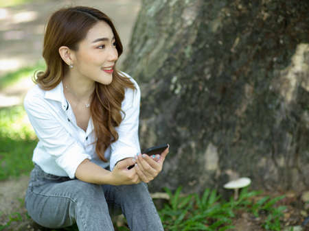 A beautiful Asian woman relaxes and sits under a tree in a garden. listen music on smartphoneの写真素材