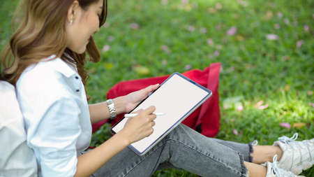 Charming female sits in a park working or sketching picture on digital tablet in a summer day. Tablet blank screen mockupの写真素材