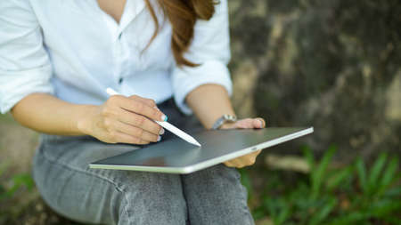 Cropped image of a female in a casual outfit working on a digital tablet while sitting under a tree in the park.の写真素材