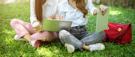 Cropped image Two female college students, relaxed and using a laptop and tablet, sit on the grass in a park and enjoy talking.の写真素材