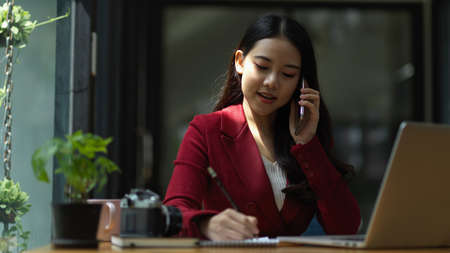 Young female manager consulting on phone and writing notes on notepads, using smart mobile phone at working placeの写真素材