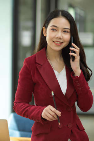 Portrait of Positive business woman in red suit talking on modern smart mobile phone in officeの写真素材