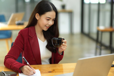 Business woman working in the office. taking notes while looking down on laptop and holding a coffee cup. Busy businesswomanの写真素材