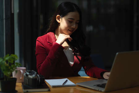 Cheerful business woman sits in office working on laptop computer, online webinar on laptop, reading business trends on website.の写真素材