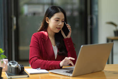 Businesswoman talking on cellphone while working on laptop computer in office. business contactの写真素材
