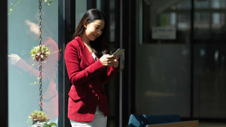 Businesswoman in red suit leaning on glass window while reading or texting message on smart mobile phone in officeの写真素材