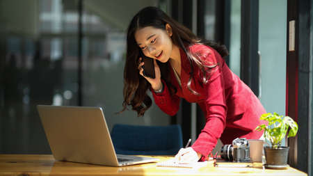 Businesswoman on a phone call. takes orders from business customer, looks on laptop and writes notes on paper while talking on phoneの写真素材