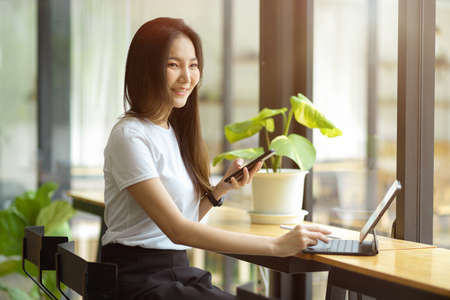 Cheerful young millennial female in casual outfits sits in co-working space at coffee shop, working on portable tablet and mobile phone.の写真素材