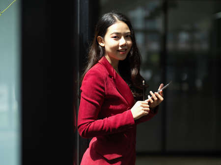 Success millennial businesswoman smile to camera, hold modern smart phone and stand in a office workplace. daylightの写真素材