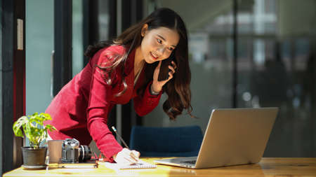 Beautiful businesswoman taking notes while having business talk on the phone with business client. Startup business concept.の写真素材