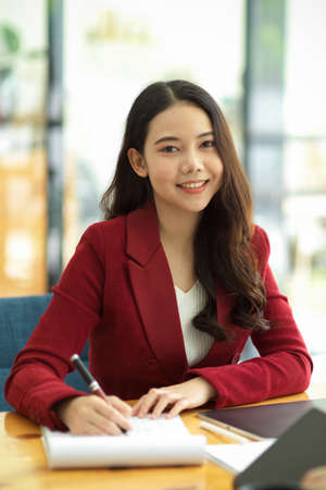 Portrait of Cheerful and happy asian businesswoman in red suit working on her business paperworks at her desk in the officeの写真素材