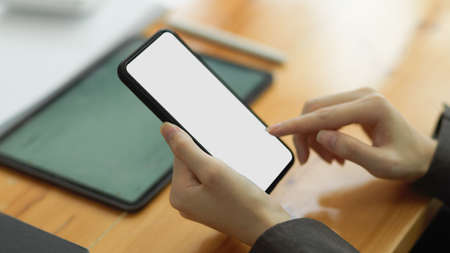 Close-up image of Female tapping finger on smartphone screen. mobile phone blank screen mockup, working desk in backgroundの写真素材