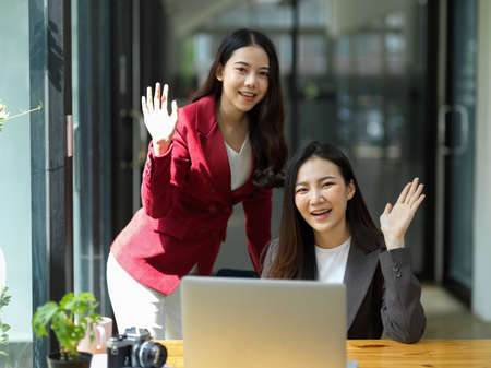 Two happy friendly businesswomen hand greeting gesture to camera in the office. Hi, hello gesture, waving with hand.の写真素材