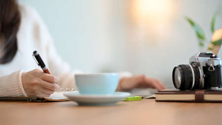 Close-up Young female in white sweater writing something on her diary, coffee cup and retro camera on wood table in a bright cafe.の写真素材