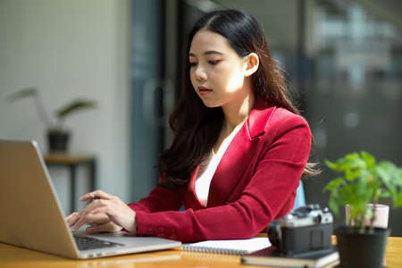 Young asian businesswoman sits at her desk in a  bright modern office workspace, working on laptop computer.の写真素材