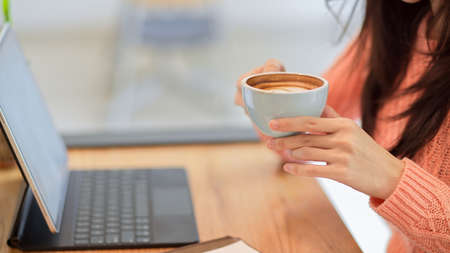 Cropped closeup image of Young female in pink sweater drinking, holding hot coffee cup while working on tablet at the cafeの写真素材