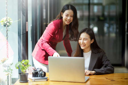 Two female colleagues using laptop computer together, discussing online project, looking at device screen together in a modern officeの写真素材