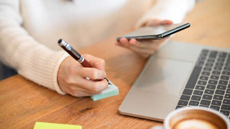 Cropped image A businesswoman in a relaxed look sits at her desk, jotting down marketing ideas on sticky notes while looking on smartphone.の写真素材
