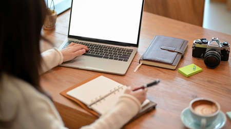 Young woman blogger, freelance photographer working on laptop computer while taking some notes on personal diary. laptop blank screen mockupの写真素材