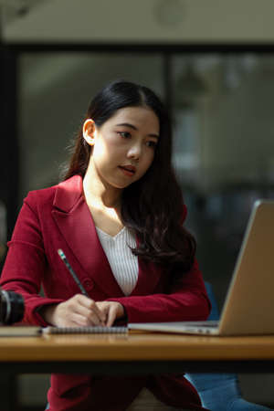 Focused young businesswoman using laptop computer and doing some paperwork or taking importance notes on notepads in the office.の写真素材