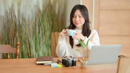 Young woman sitting in a beautiful coffee shop, drinking hot coffee in the morning, take a break after work on laptop computer.の写真素材