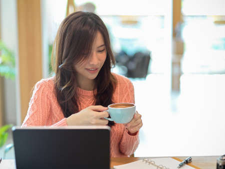 Beautiful young woman in pink sweater drinking her coffee in the cafe while working, online studying on her portable tablet computer.の写真素材
