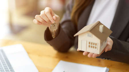 Cropped image of Young female house insurance broker or real estate agent holding house key and house model at the desk.の写真素材