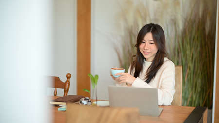 Charming young woman working on her laptop computer while sipping warm coffee at Japanese cafe style.の写真素材