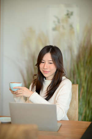 Beautiful millennial asian woman drinking hot coffee to warm herself while working on her laptop computer at cafe.の写真素材