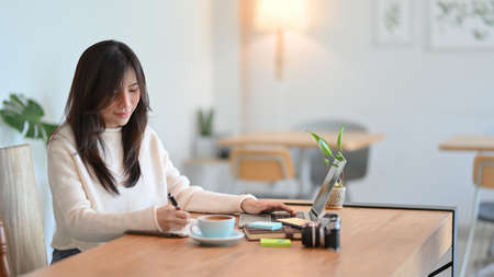 Pleased young asian female making a notes on her diary while working on laptop computer at minimalist cafeの写真素材