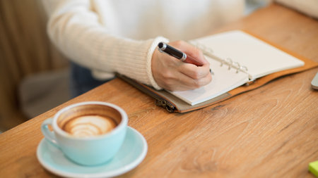 Cropped closeup image of Female in white sweater writing or taking notes something on her notebook, tasty coffee on the table.の写真素材