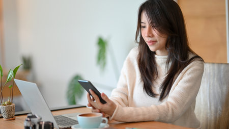 A lovely Asian young lady sits at the cafe co-working space, reading online business news on her smart phone.の写真素材