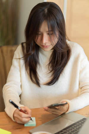 Portrait of young asian female taking notes on memo pads while looking on mobile screen, working from home.の写真素材