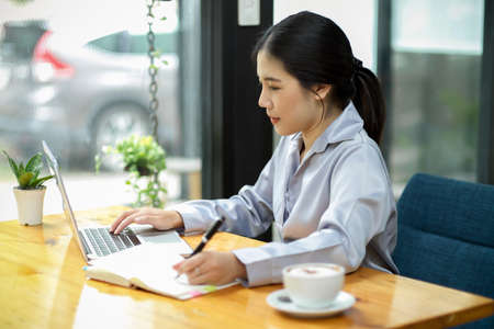 Side view of asian female secretary working and looking at laptop screen at her desk, writing and managing her boss schedule on notebook.の写真素材
