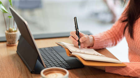 Close-up Pretty female writing her works on her personal schedule book while working on tablet computer.の写真素材
