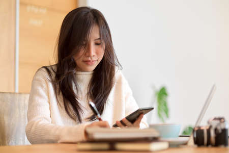 Beautiful college student writing on a memo notes, taking notes while looking at mobile screen.の写真素材