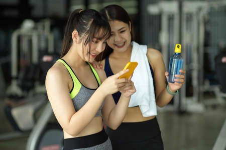 Two of sporty girl standing in fitness studio looking on smartphone screen together after finished...
