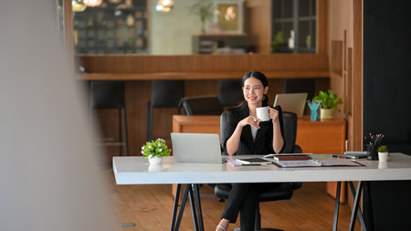 Elegant businesswoman in a black suit sits at her desk, sipping coffee and pondering her thoughts as she gazes out the window.の写真素材