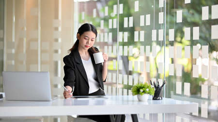 A smart confident young asian business woman drinking coffee during working on tablet computer in the modern office.の写真素材