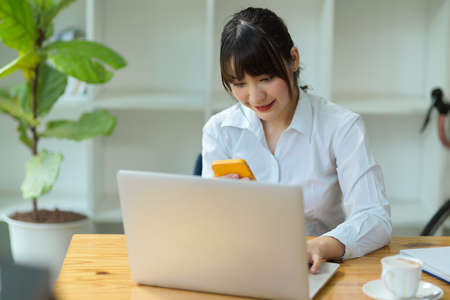 Female college student using smartphone for searching an informations while doing assignment on laptop.の写真素材