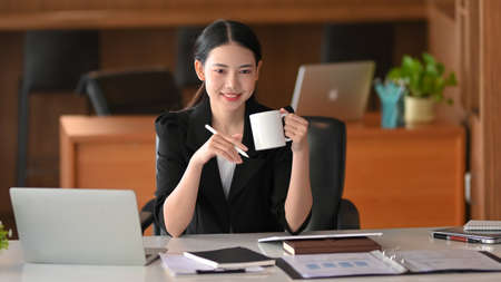 Beautiful and appealing business female boss drinks coffee while working on her laptop and reports.の写真素材