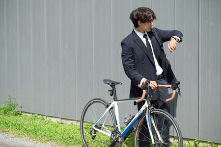 Asian young businessman walking outdoors and pushing a bicycle while looking at his wristwatch.の写真素材