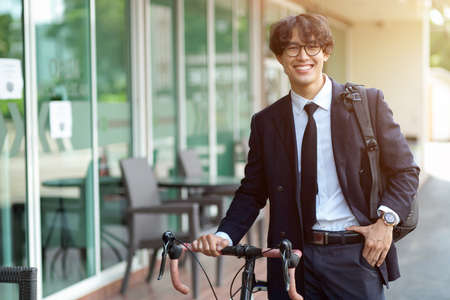 Confident handsome asian young businessman going to work by bike, walking outdoors with a bicycle in a modern street.の写真素材