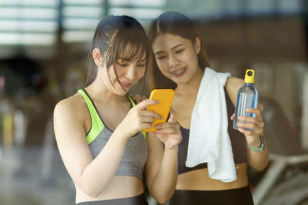 Two asian female at the gym, taking rest from over exercising and looking something on smartphone.の写真素材