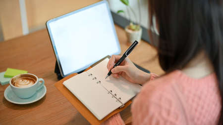 Close-up Young female hold notebook and write something on paper while looking at tablet screen.の写真素材